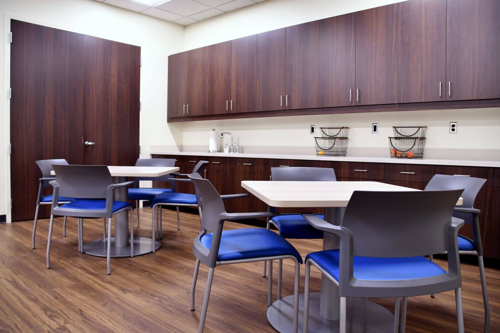 Office breakroom with dark wood cabinets, square dining tables, and blue and grey chairs