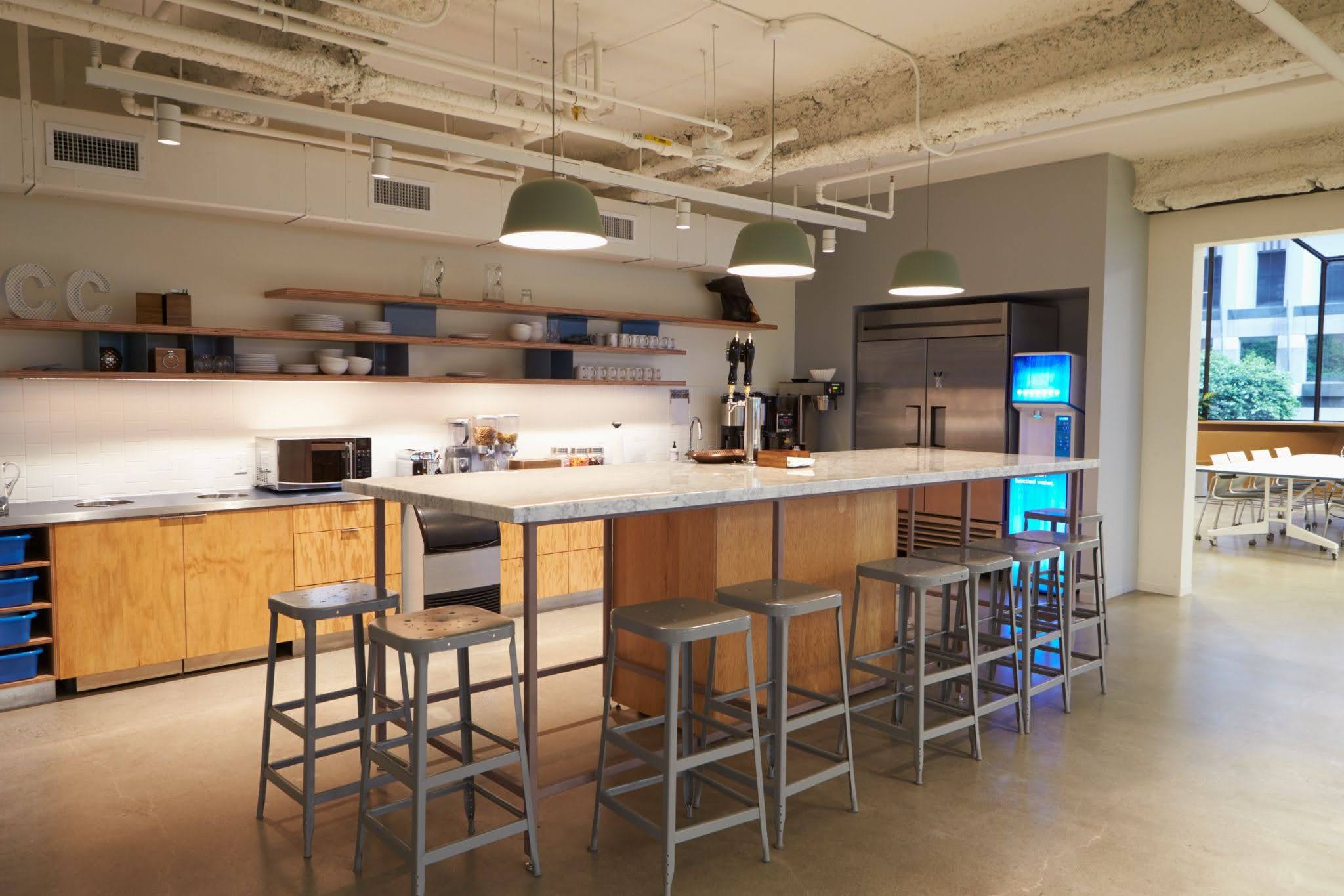 Modern office breakroom featuring a wooden kitchen counter, marble-topped island with grey bar stools, and pendant lighting.