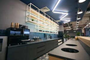 Modern office kitchen breakroom with coffee machine, blue tile backsplash, and hanging metal shelving unit